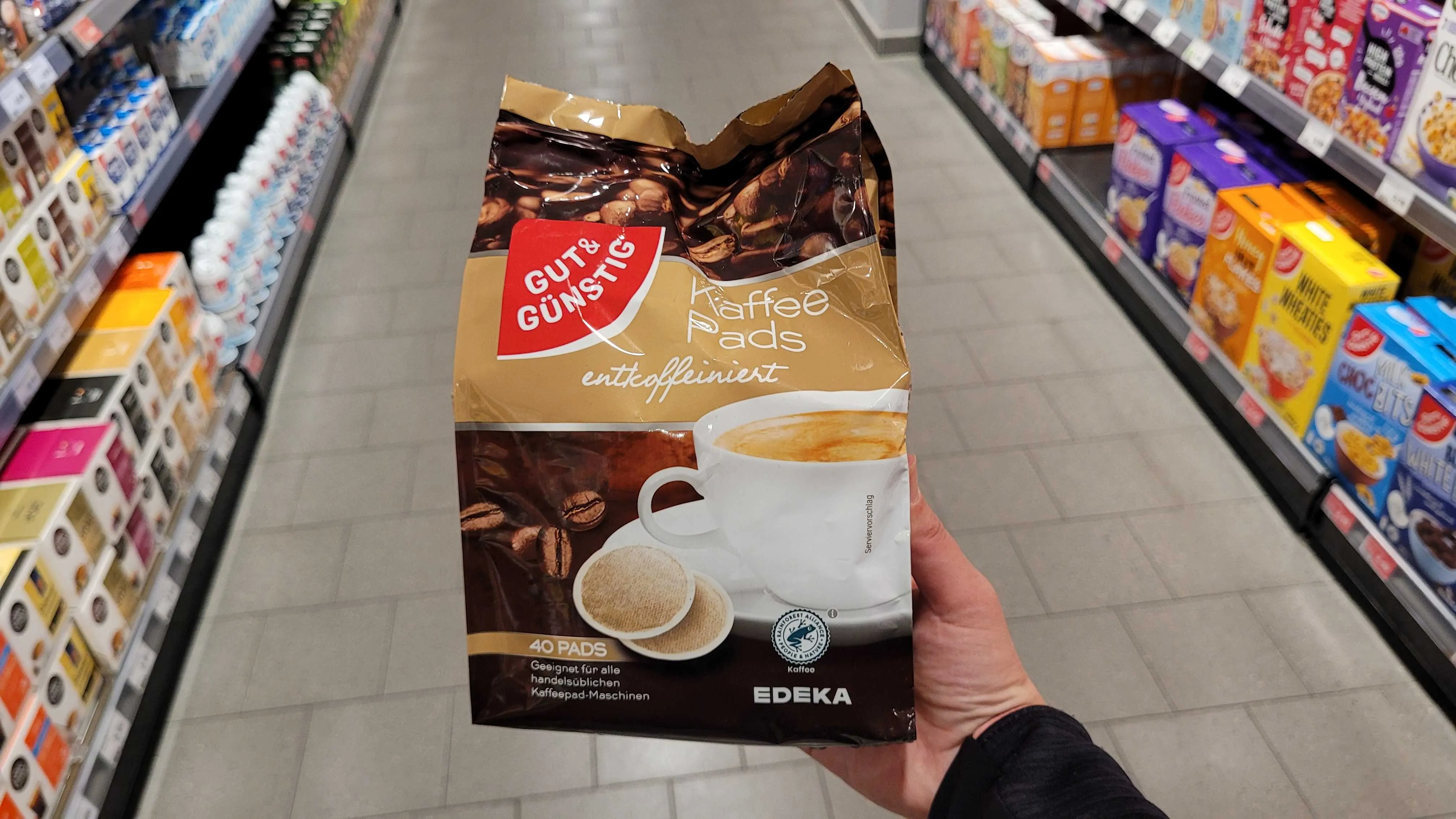 Kaffeepads entkoffeiniert im Test: Eine Hand hält eine Packung über einem Fliesengang.
