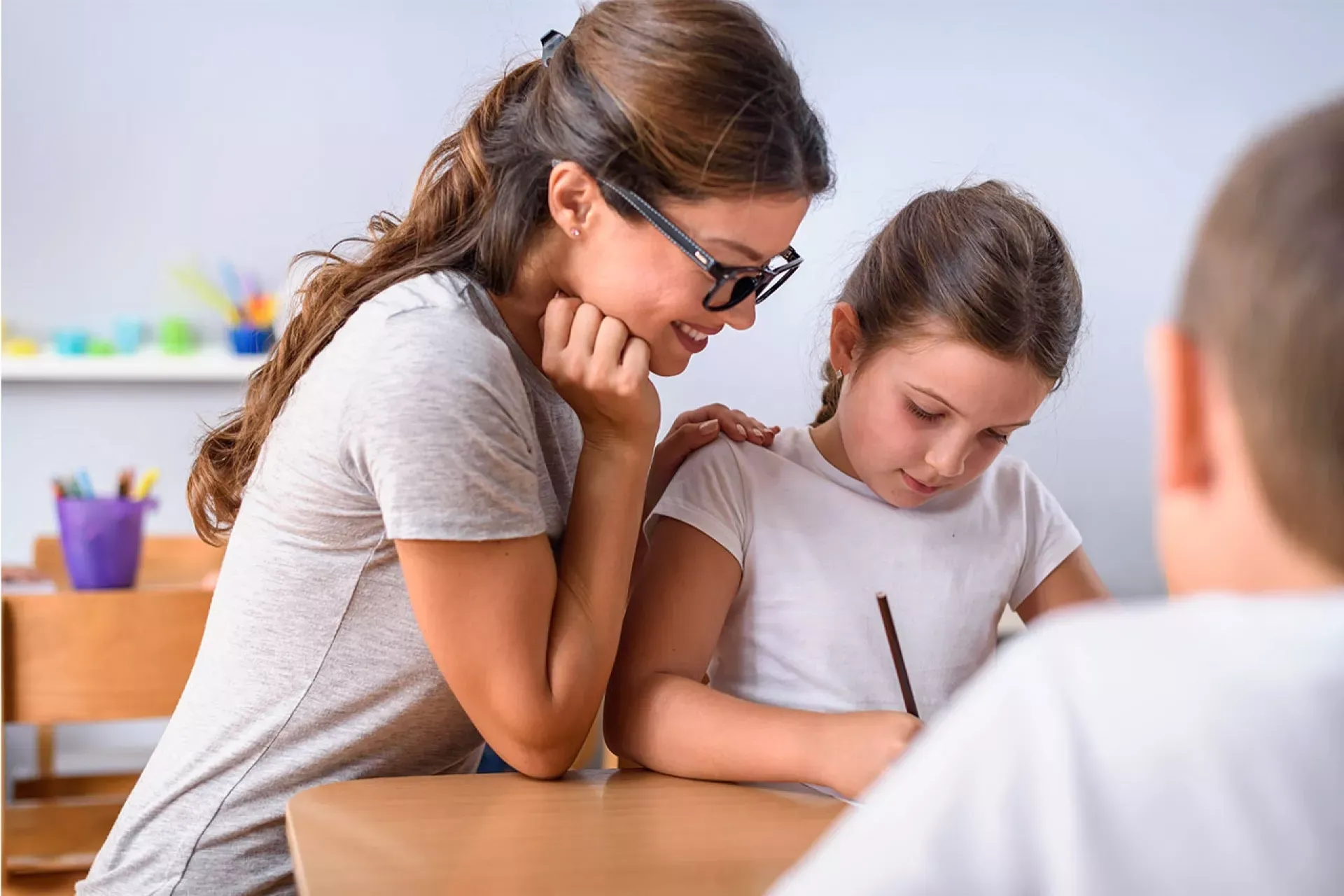 Lehrerin sitzt mit 2 Kindern am Tisch und unterstützt ein Kind beim Schreiben
