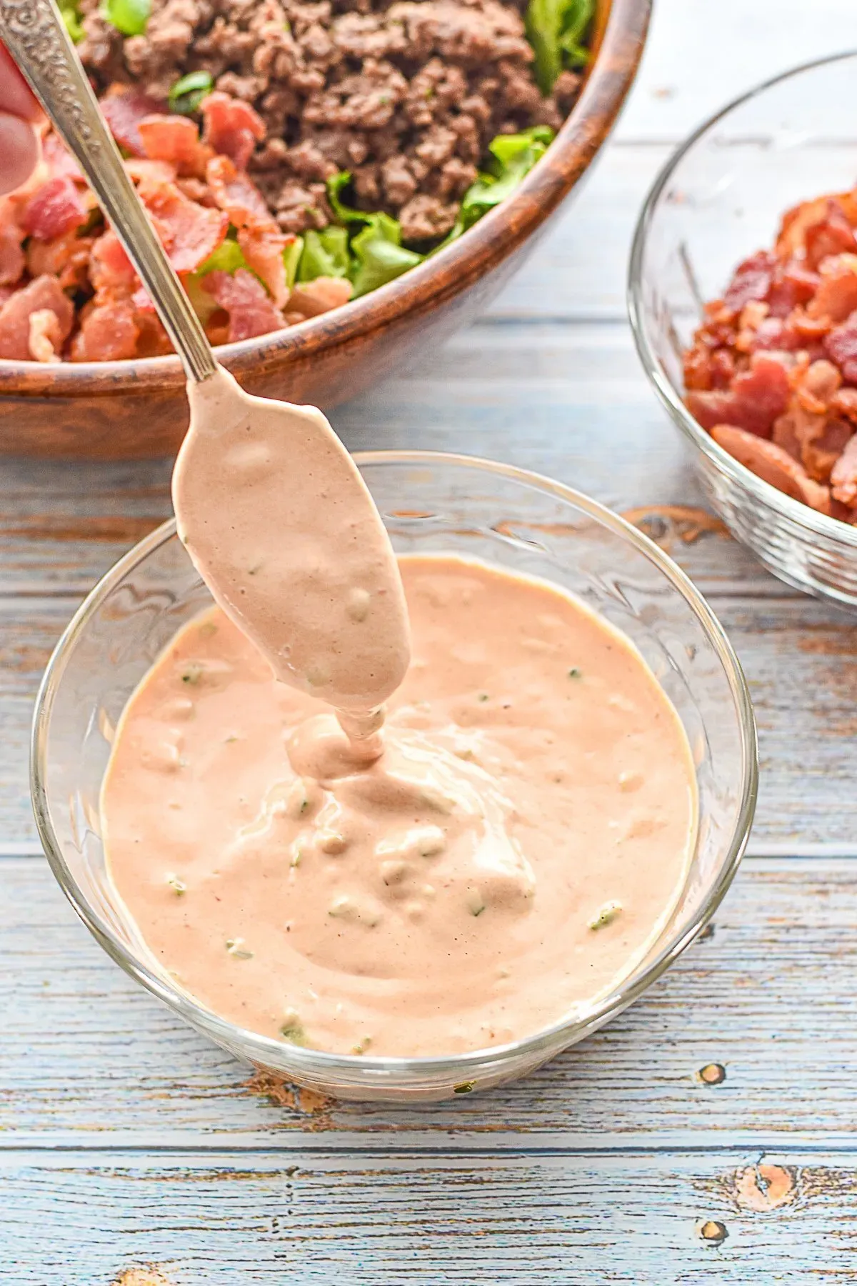 low fodmap burger sauce drizzling from a spoon into a clear bowl in front of a burger bowl and bowl of bacon pieces