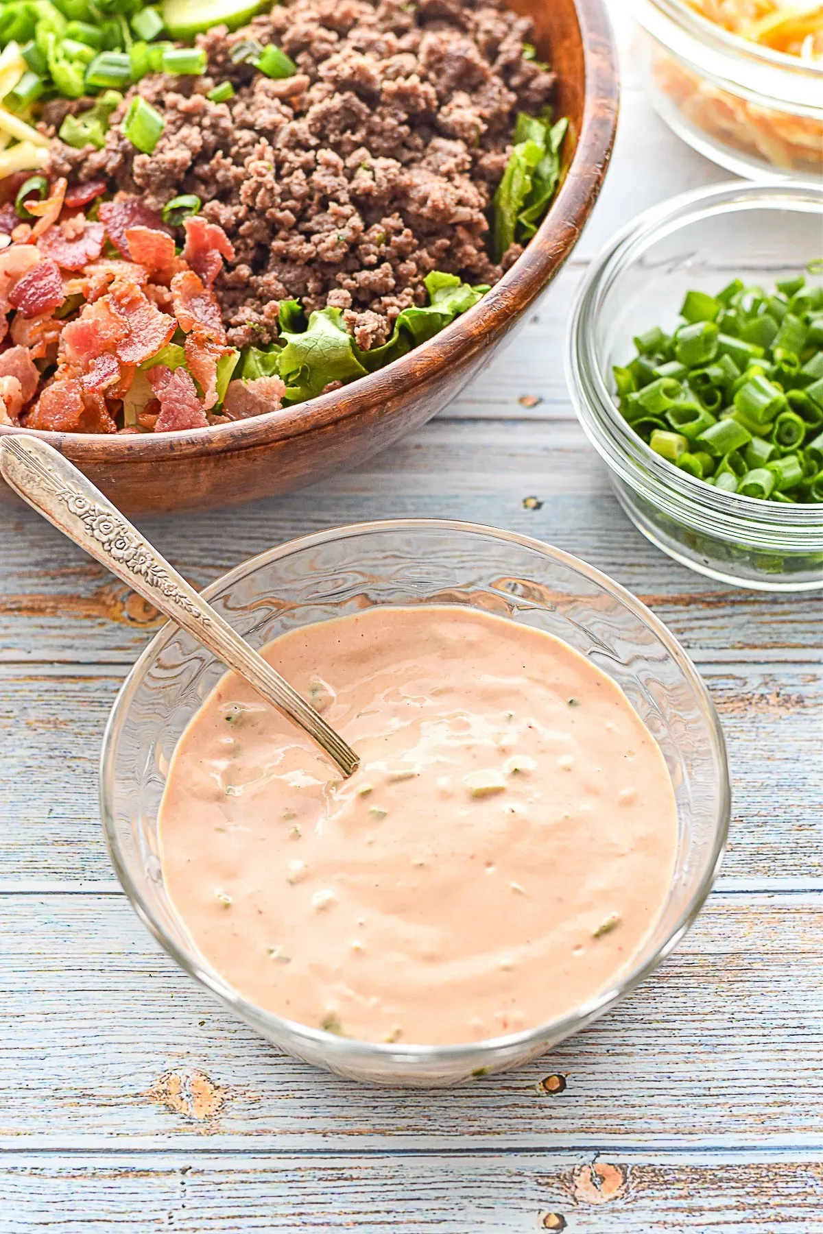 low fodmap burger sauce in a glass bowl with a spoon in front of a burger bowl and bowls of chopped scallion greens and shredded cheese
