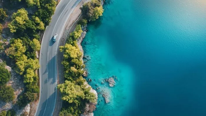 Luftaufnahme einer Küstenstraße mit einem fahrenden Auto neben blauem Meer und grünen Bäumen.