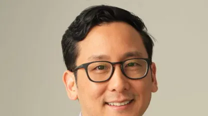 Mann mit kurzen schwarzen Haaren und Brille, lächelnd vor schlichtem Hintergrund