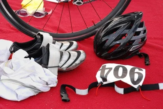 Triathlon-Ausrüstung, bestehend aus einem Fahrrad, einem Helm, Schuhen, einer Trikot und einer Startnummer auf rotem Untergrund.