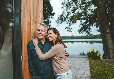 Umschlungen blickt ein älteres Paar in das Fenster ihres Hauses.