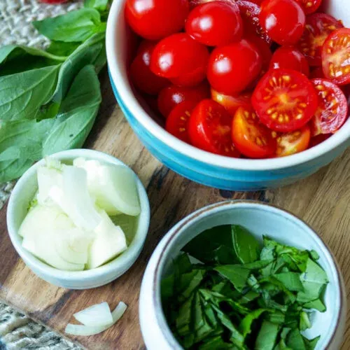 Vorbereitete Zutaten wie Zwiebeln, Basilikum und halbierte Tomaten auf einem Holzbrett.