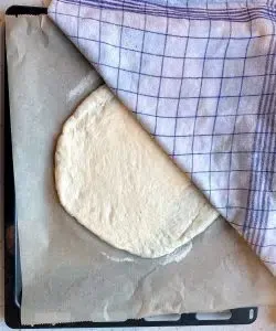 Zwei ausgerollte Fladenbrote liegen auf einem mit Grieß bestreuten Backblech, bedeckt mit einem feuchten Tuch